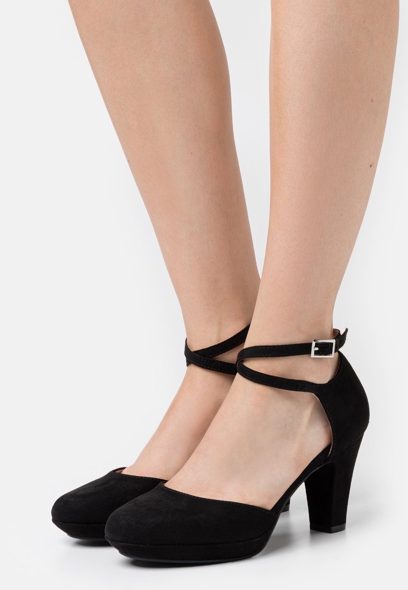 Pieds portant des chaussures à talons hauts en daim noir avec des brides à la cheville et des boucles argentées, présentés sur un fond blanc uni.