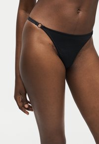 Κοντινό πλάνο ενός ατόμου που φορά μαύρο κάτω μέρος μπικίνι με λεπτομέρεια κρίκου στην αριστερή πλευρά, που στέκεται μπροστά σε απλό ανοιχτό φόντο.