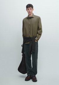Camicia verde oliva a maniche lunghe con bottoni, maglione nero legato in vita, jeans scuri, scarpe di pelle marrone, con una borsa marrone. Texture liscia.