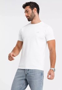 Hvid bomulds t-shirt med korte ærmer, der har et subtilt logo på brystet. Tætsiddende design, glat tekstur, kombineret med lyseblå jeans.