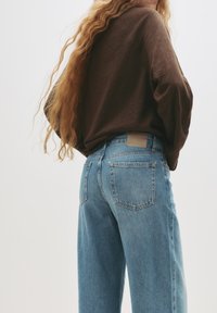 Modre kavbojske hlače sproščenega kroja, s svetlim pranjem, zadnjimi žepi in svetlo rjavim označevalnim trakom na pasu. V kompletu z rjavim dolgim rokavom.