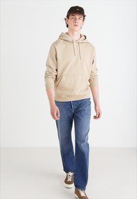 Sweatshirt beige à capuche avec une poche kangourou, associé à un jean droit bleu et des baskets marron avec des semelles blanches, marchant vers l'avant.