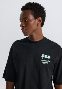 Svart bomullströja med avslappnad passform, med en mintgrön grafik på vänstra bröstet som inkluderar stiliserade stolar och text.