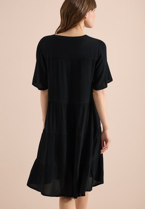 Mujer vestida con un vestido negro hasta la rodilla con mangas cortas, vista desde atrás sobre un fondo beige liso.