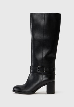 Botte en cuir noire montant jusqu'aux genoux avec talon carré et bride décorative à boucle autour de la cheville, vue de profil sur un fond uni.