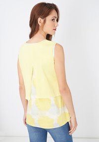 Top sin mangas con un frente amarillo sólido, diseño en capas y tela blanca con patrones de salpicaduras amarillas en la parte inferior.