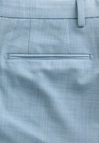 Tissu bleu clair en gros plan montrant une poche arrière avec une couture passepoilée horizontale et une partie d’une boucle de ceinture sur un pantalon sur mesure.