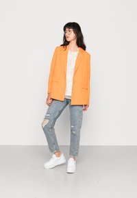 Blazer oversized laranja com lapelas recortadas, combinado com uma blusa branca estampada, jeans claros rasgados e ténis brancos robustos.