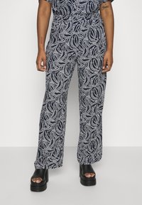 Námořnické modré široké kalhoty s bílým točivým vzorem, vyrobené z lehké tkaniny. Mají elastický pas a jsou nošeny s černými platformovými sandály.