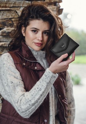 Black leather wallet with curved flap and embossed logo, held by a person wearing a knitted cream sweater and maroon vest. Textured finish.