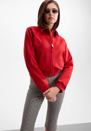 Jeune femme portant des lunettes de soleil noires, une veste rouge zippée et un pantalon à carreaux noir et blanc, debout avec les mains jointes.