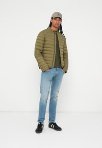 Chaqueta de plumas verde oliva con costuras verticales, camisa verde oscura, jeans azul claro y zapatillas negras con rayas blancas. Gorra gris.