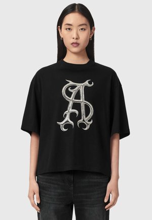 Ung kvinde med langt sort hår bærer en løs sort t-shirt med et stort udsmykket sølv "AS" monogram foran.