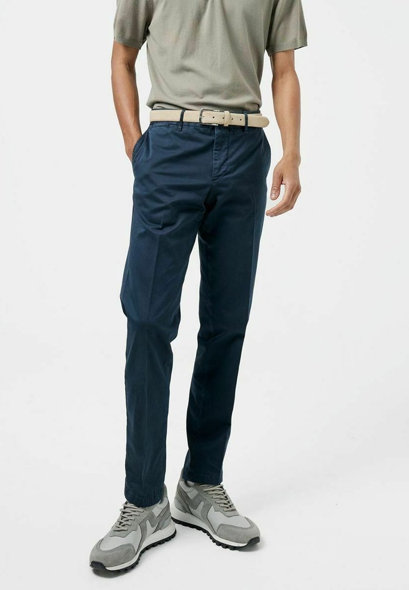 Homme portant un pantalon bleu marine, une ceinture beige, une chemise à manches courtes gris-vert et des baskets grises, debout les mains détendues.