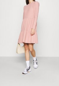 Abito lungo a maniche lunghe rosa a coste con gonna a strati, abbinato a calze bianche e sneakers. Una borsa crema è tenuta nella mano sinistra.