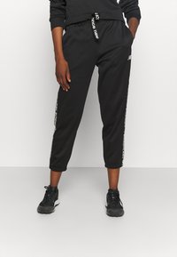 Svarta joggingsbyxor med elastisk midja och dragsko. Har vitt logodetalj på sidan och en avslappnad, kort passform. Bärs med svarta sneakers.