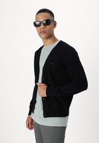 Crna cardigan jakna s V-izrezom, prednjim dugmadi i malim logotipom s lijeve strane, složena preko svijetlosive majice. Glatka tekstura.