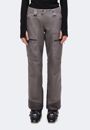 Personne portant un pantalon de neige gris avec des poches zippées sur les cuisses et des chaussures de ski noires, debout devant un fond uni.