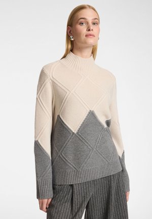 Vrouw draagt een trui met hoge hals en grote ruitpatronen in beige en grijs gebreide stof, gecombineerd met een grijze ribbroek.