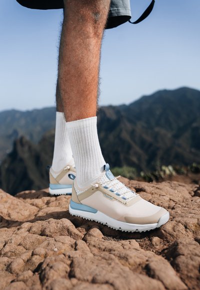 Lightweight hiking shoes in beige and light blue, with mesh panels, rubber soles, and a flexible structure. Paired with white crew socks.