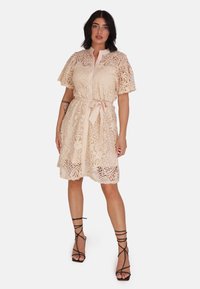 Sukienka koronkowa w jasnym beżu, z krótkimi falbaniastymi rękawami, talią zdobioną paskiem oraz skomplikowanymi wzorami kwiatowymi, z przodem zapinanym na guziki.
