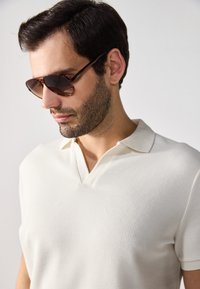 Kurzärmliges, cremefarbenes Polohemd aus strukturiertem Stoff mit V-Ausschnittkragen. Braungetönte Sonnenbrillen runden den Look ab.