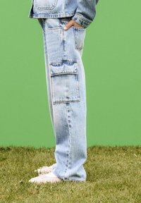 Pantaloni cargo in denim azzurro chiaro con tasche laterali, vestibilità leggermente rilassata e cuciture visibili. Le scarpe sono di colore rosa chiaro con lacci, su erba.