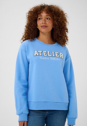 Vrouw met krullend haar draagt een lichtblauwe trui met de tekst "ATELIER Creative Collective" en blauwe jeans tegen een effen achtergrond.