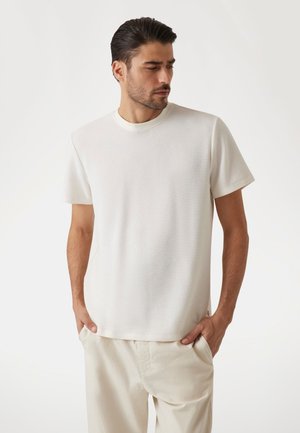 White short-sleeve t-shirt made of textured fabric, with a round neck and slightly loose fit. Paired with light-colored pants.
