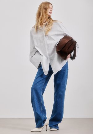 Kobieta w za dużej, jasnej koszuli w paski, niebieskich szerokich dżinsach, białych szpilkach, trzymająca brązową torebkę, stojąca na jednolitym tle.