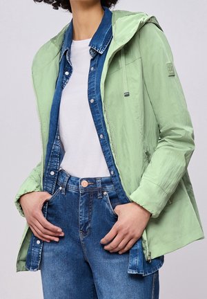 Person iført en lysegrøn hættetrøje over en blå denimskjorte og hvid t-shirt, med hænderne i lommen på blå denimjeans.