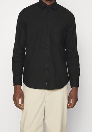 Mann trägt ein langärmeliges schwarzes Hemd mit Knöpfen und Brusttasche sowie beige Hose und steht vor einem einfarbigen Hintergrund.