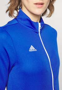 Blå zip-up treningsjakke med lette teksturer, med et hvitt Adidas-logo på brystet og høy hals.