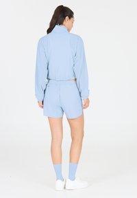 Helles blaues Sportoutfit bestehend aus einer Langarmjacke mit hohem Kragen und perforierten Seiteneinsätzen, kombiniert mit passenden Shorts und weißen Turnschuhen.