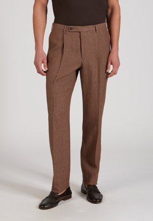 Hombre con pantalones marrones ajustados y texturizados, camisa negra y mocasines de cuero marrón oscuro, de pie frente a un fondo neutro.