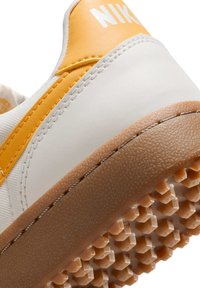 Valkoinen ja oranssi lenkkari, jossa on sileä synteettinen päällyste, teksturoitu kumipohja, jossa on kuusikulmainen kuvio ja hallitseva logotunnus.