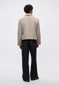 Szara kurtka z mieszanki wełny z wyciętym kołnierzem, krótkim krojem i bocznymi kieszeniami, zestawiona z ciemnoniebieskimi spodniami o szerokich nogawkach z delikatnym przeszyciem.