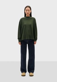 Pull oversized vert foncé à manches longues, col haut, associé à un jean en denim foncé à jambes larges et des sabots beige. Texture lisse.