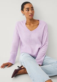 Pull en maille violet clair avec un col en V, des poignets et un ourlet côtelés. Associé à un jean déchiré bleu clair et des talons bordeaux.
