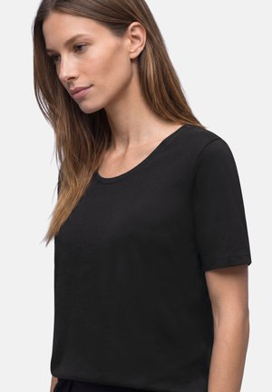 Woman with long brown hair wearing a plain black short-sleeve t-shirt, facing slightly left against a light gray background.