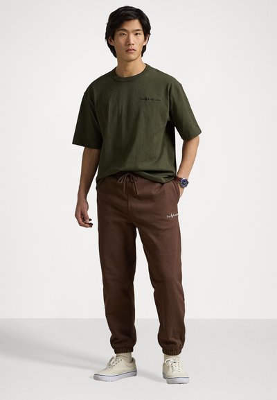 Tricou din bumbac de culoare verde măsliniu, cu mâneci scurte și un logo pe piept; pantaloni joggers căprui cu șnur și manșete la glezne; purtat cu pantofi sport deschiși la culoare.