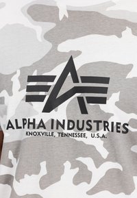 Γκρι και λευκό καμουφλάζ μπλούζα t-shirt με μαύρο λογότυπο που έχει την ένδειξη "ALPHA INDUSTRIES" και κείμενο τοποθεσίας εκτυπωμένο κάτω από αυτό.