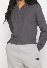 Donna che indossa una felpa grigia scura con mezza zip e pantaloni morbidi grigi chiari con etichetta "Nicki Studios", mani nelle tasche.