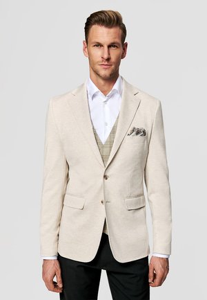 Mężczyzna w beżowej marynarce, białej koszuli, wzorzystej kamizelce i czarnych spodniach stoi na jednolitym tle.