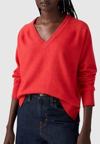 Pull rouge en V à manches longues, fait de matériau doux. Présente des poignets côtelés et une coupe décontractée, associé à un jean en denim foncé.