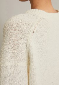 Hellbeige Strickpullover mit strukturiertem, offenem Gewebe an den Schultern und ribbeligem Halsausschnitt, der den weichen Stoff und das lässige Design betont.