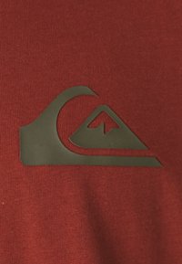 Červená látka s hladkým povrchom, na ľavej strane sa nachádza tmavozelené logo vlny a hory.