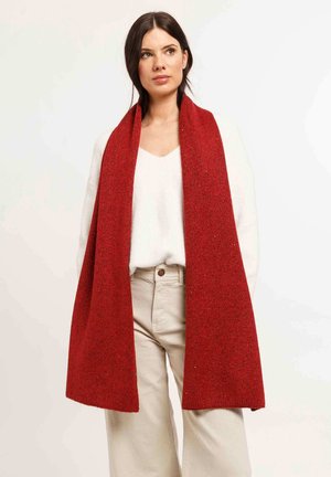 Woman wearing a long red scarf over a white sweater and beige pants, standing against a plain light background.