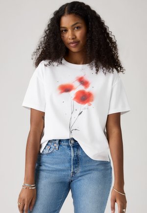 Mladá žena s kudrnatými vlasy, která má na sobě bílé tričko s potiskem červených máků a modré džíny, stojí před jednobarevným pozadím.