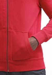 Personne portant un sweat à capuche rouge zippé, une main partiellement dans la poche avant, sur fond blanc.